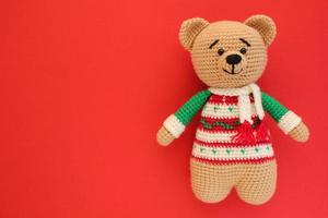 Crochet amigurumi handmade stuffed soft teddy bear toy in colored sweater on red background. Handwork, hobby. Craft diy newborn pregnancy concept. Knitted doll for little baby. Closeup flat lay out photo