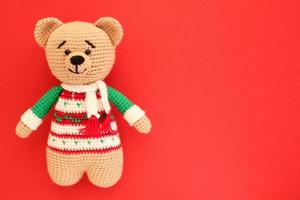 Crochet amigurumi handmade stuffed soft teddy bear toy in colored sweater on red background. Handwork, hobby. Craft diy newborn pregnancy concept. Knitted doll for little baby. Closeup flat lay out photo