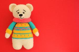 Crochet amigurumi handmade stuffed soft teddy bear toy in colored sweater on red background. Handwork, hobby. Craft diy newborn pregnancy concept. Knitted doll for little baby. Closeup flat lay out photo