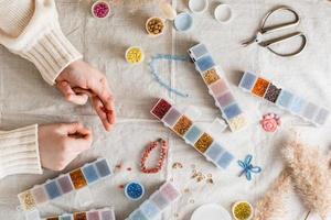 The girl's hands are weaving a beaded bracelet and items for beading on the table. Development of creative skills and fine motor skills for children. Top view. photo