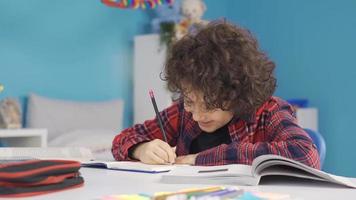 Primary school student doing homework at the desk is studying with pleasure. Cute curly haired male student doing homework, taking notes, looking at book. video