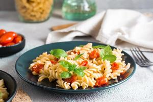 A plate of pasta with tomatoes and basil on a plate and cooking ingredients on the table. Mediterranean cuisine.