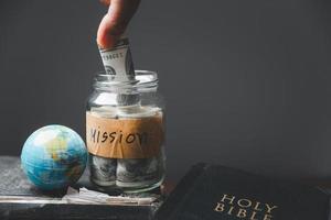 Saving jars full of money and globe with Holy Bible for mission, Mission christian idea. Hand holding dollar with bible on wooden table, Christian background for great commission or earth day concept. photo