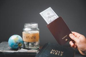 ahorro frascos lleno de dinero y globo con santo Biblia para misión, misión cristiano idea. mano participación dólar y pasaporte en mesa, cristiano antecedentes para genial comisión o tierra día concepto. foto