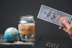 Saving jars full of money and globe with Holy Bible for mission, Mission christian idea. Hand holding dollar with bible on wooden table, Christian background for great commission or earth day concept. photo