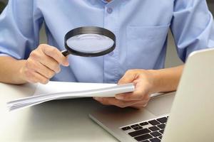 Close up hand of businessperson looking at documents through magnifying glass and laptop in front. Concept of business and data validation. photo