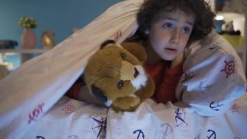 schattig jongen schuilplaats onder de bed met zijn speelgoed- hond, spelen. een jongen spelen verbergen en zoeken verbergt onder de deken met zijn pluche speelgoed- en is gelukkig. video