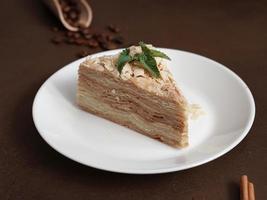 Delicious Napoleon Cake with cream on a white plate decorated with a sprig of mint on a Brown Background. A cinnamon stick, badyan, coffee beans on a brown background. Copy space photo