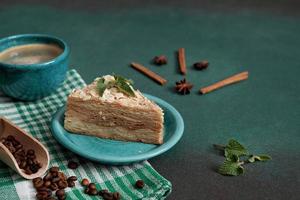 delicioso Napoleón pastel con crema en un turquesa plato decorado con un puntilla de menta en un verde antecedentes. un taza de caliente café, canela palo, badyan, café frijoles en un verde antecedentes foto