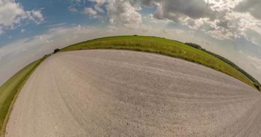 tiny planet transformation with curvature of space among fields on gravel road in sunny day with sky and fluffy clouds video