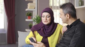 musulmán familia mira a el teléfono a hogar. mujer en hijab y su marido son sentado en el sofá y mirando a alguna cosa en el teléfono. video