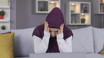 muçulmano mulher dentro hijab triste, obtendo mau notícias, leitura, ouvindo. muçulmano mulher sentado em sofá às lar, olhando às computador portátil e sentindo-me triste. video