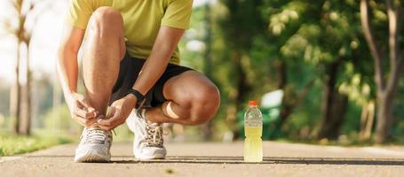 Young athlete man tying running shoes with Energy Drink water, male runner ready for jogging outside, asian Fitness walking and exercise in the park morning. wellness, wellbeing and sport concepts photo