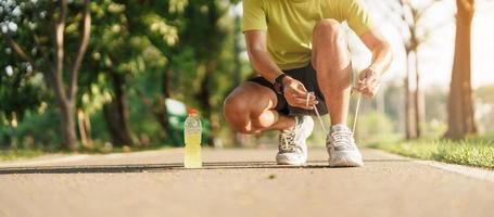 Young athlete man tying running shoes with Energy Drink water, male runner ready for jogging outside, asian Fitness walking and exercise in the park morning. wellness, wellbeing and sport concepts photo