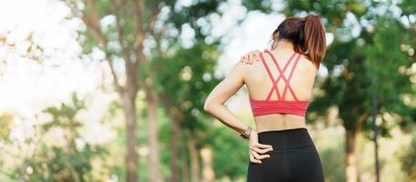 Young fitness woman holding her sports injury shoulder and neck, muscle painful during training. Asian runner female having body problem after exercise outside in summer photo