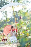 Young Asian woman wearing a white dress poses with a rose in rose garden photo