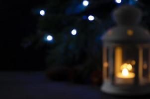 Blurred picture for background. White lantern with a burning candle, pine branch and garland lights on background. Christmas mystery mood. photo