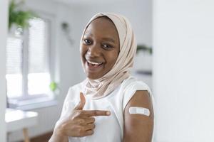retrato de un musulmán africano hembra sonriente después consiguiendo un vacuna. mujer participación abajo su camisa manga y demostración su brazo con vendaje después recepción vacunación. foto