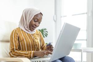mujer musulmana elegante y atractiva que usa una computadora portátil móvil que busca información de compras en línea en la sala de estar en casa. retrato de mujer feliz comprando productos a través de compras en línea. pagar con tarjeta de crédito