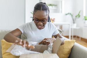 Young satisfied happy african girl woman lady shopaholic customer sit on sofa unpack parcel delivery box, online shopping shipment concept. photo