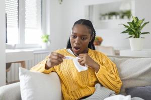 Ill african young woman covered with blanket blowing running nose got fever caught cold sneezing in tissue sit on sofa, sick allergic black girl having allergy symptoms coughing at home, flu concept photo