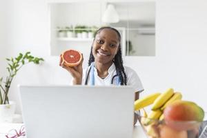 Portrait of young smiling female nutritionist in the consultation room. Nutritionist desk with healthy fruit, juice and measuring tape. Dietitian working on diet plan. photo