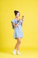 imagen de hermosa asiático niña en pie en amarillo antecedentes y utilizando teléfono foto