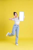 full body image of young Asian girl holding phone with cheerful face on yellow background photo