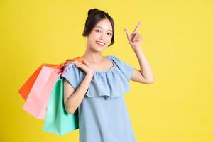 imagen de un hermosa asiático niña en un vestir participación un compras bolso en su mano posando en un amarillo antecedentes foto