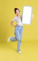 full body image of young Asian girl holding phone with cheerful face on yellow background photo