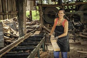 mujer sonrisas y guiños uno ojo mirando a cámara, cargando un tablero en un aserradero. foto