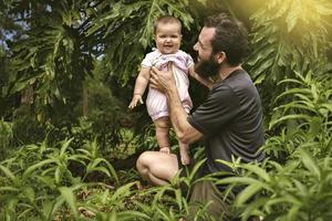 un bebé niña mira a el cámara sonriente, su padre sostiene al aire libre vida foto