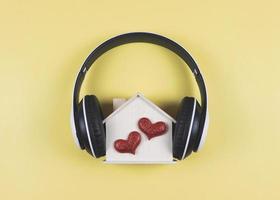 flat lay of wooden model house with two red glitter hearts covered with headphones isolated on yellow background. music or podcast, home of love, valentines. photo