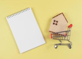 flat layout of wooden house model in shopping trolley or shopping cart with blank page opened notebook on yellow  background, home purchase concept.