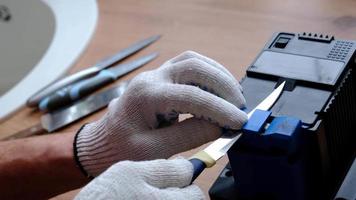 Schärfung ein Messer auf ein elektrisch Anspitzer beim heim. das des Mannes Hand fährt das Messer Klinge zwischen das Blau Spitzer, Staub fliegt auf das Maschine. 4k, schleppend Bewegung video