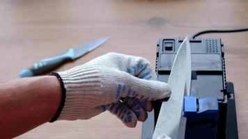 verscherping een mes Aan een elektrisch slijper Bij huis. de man's hand- schijven de mes blad tussen de blauw slijpers, stof vliegt Aan de machine. 4k, langzaam beweging video