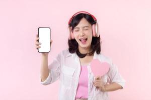contento alegre sonriente asiático mujer con mano participación corazón y auriculares demostración blanco pantalla móvil teléfono o nuevo teléfono inteligente música solicitud anuncio Bosquejo aislado en rosado estudio antecedentes. foto