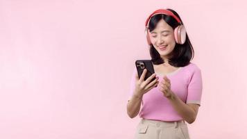 Portrait cheerful young asian woman enjoy listening audio by smartphone music application against pink. Happy smiling female person with headphone. Sound, leisure, lifestyle, technology concept
