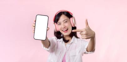 Happy cheerful smiling asian woman with wireless earphones showing blank screen mobile phone or new smartphone music application advertisement mockup isolated on pink studio background.