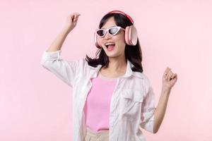 happy young asian woman model with stylish trendy sun glasses enjoy listening music by headphone audio and dancing isolated on pink studio background. technology, girl fashion, accessory concept. photo