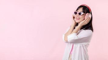 happy young asian woman model with stylish trendy sun glasses enjoy listening music by headphone audio and dancing isolated on pink studio background. technology, girl fashion, accessory concept. photo