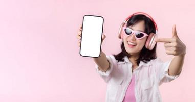 Happy cheerful smiling asian woman with wireless earphones showing blank screen mobile phone or new smartphone music application advertisement mockup isolated on pink studio background. photo