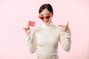 Fascinating fun joyful young woman of Asian ethnicity 20s years old with wear sunglasses wears white shirt hold in hand credit bank card isolated on plain pastel light pink background studio portrait. photo