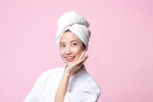 hermosa joven asiático mujer contento con limpiar cara piel en toalla y bata de baño, spa traje en rosado antecedentes. protección de la piel, tratamiento, bienestar terapia, facial cuidado, belleza hembra salud, cosmetología concepto foto