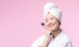 Portrait happy young asian woman with natural make up face holding cosmetic skin powder blusher isolated on pink background. Female apply skincare brush treatment. beauty product, cosmetology concept. photo