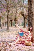 dama en tradicional kimono vestir participación libro y mirando Cereza florecer en primavera festival. foto