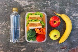 Sandwiches, fruits and vegetables in food box, water on old wooden background. Top view. Flat lay. photo