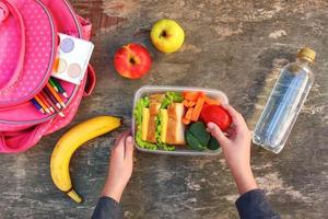 sándwiches, frutas y vegetales en comida caja, mochila en antiguo de madera antecedentes. concepto de niño comiendo a escuela. parte superior vista. plano poner. foto