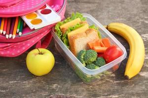 sándwiches, frutas y vegetales en comida caja, mochila en antiguo de madera antecedentes. foto