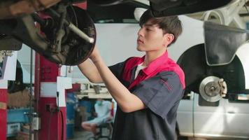 One young expert Asian male automotive mechanic technician is screwing car wheel nuts on lifting with wrench for repair at garage. Vehicle maintenance service works, industry occupation business jobs. video